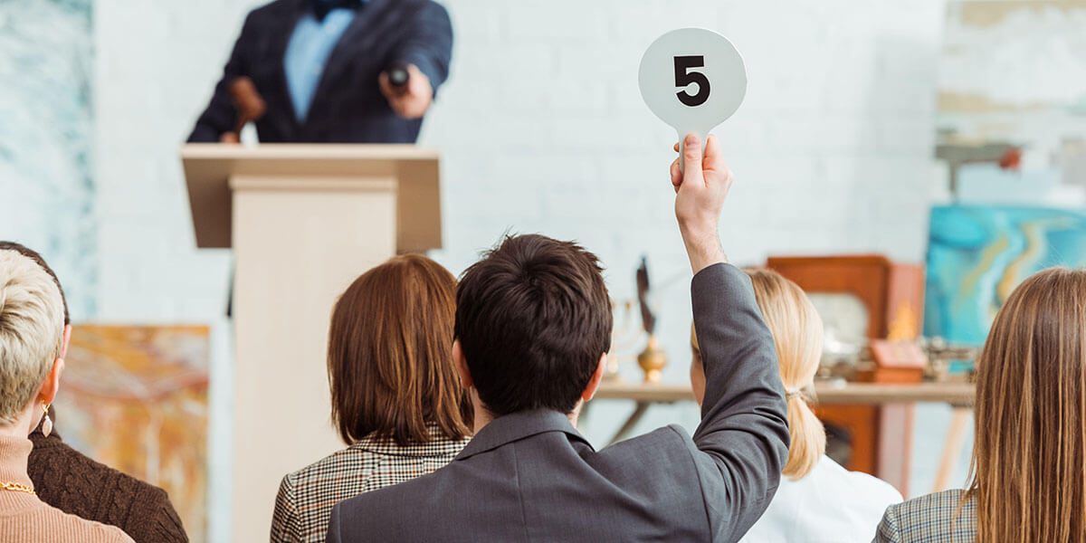 A photograph of an auction master at a podium in front of a group of people with one man raising a paddled with the number 5 on show.