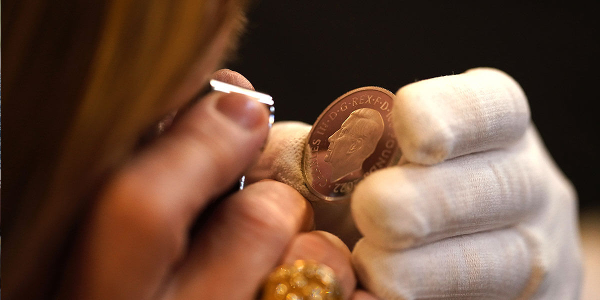 A close up of a lady inspecting a gold coin featuring the portrait of King Charles III with a small microscope.