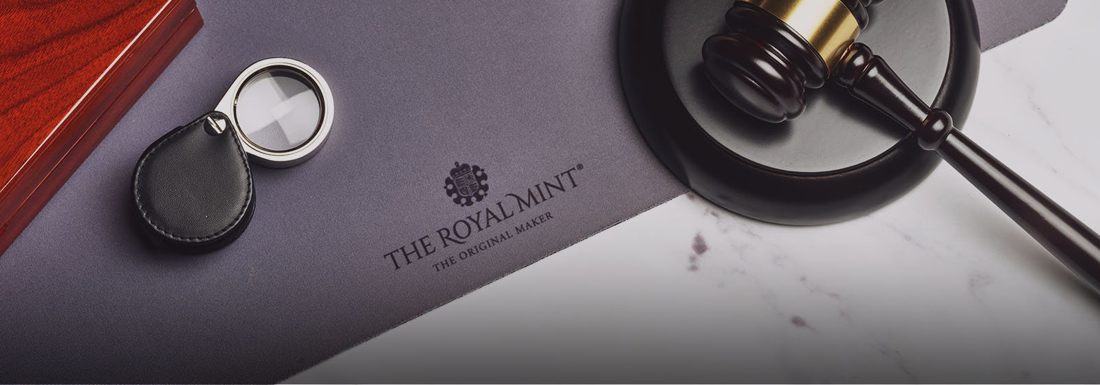 A dark wood gavel and striking block, foldable magnifying glass, mahogany coin storage box sitting on a coin mat embossed with The Royal Mint The Original Maker.